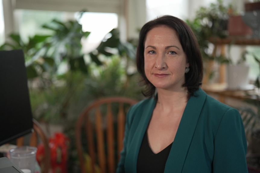 A white woman with brown hair wearing a green jacket.  She is sitting in an office filled with plants